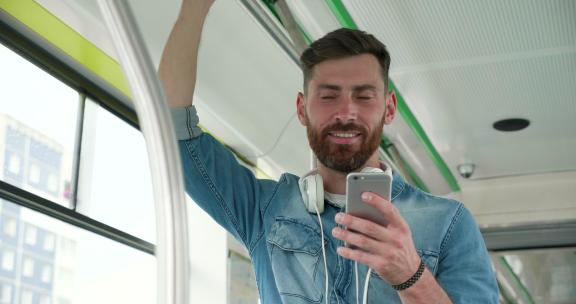 穿着休闲的迷人男人使用有轨电车，一只手拿着智能手机，另一只手扶着扶手。看起来微笑而华丽。他脸上有好心情和积极的情绪。