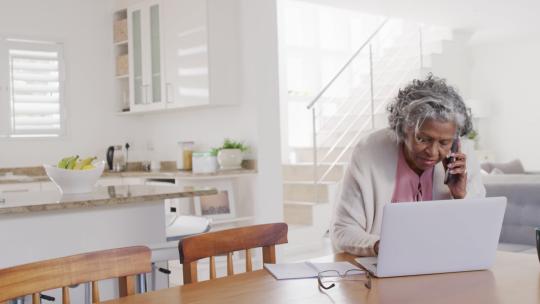 高级非裔美国妇女坐在桌旁，使用笔记本电脑和智能手机交谈的肖像