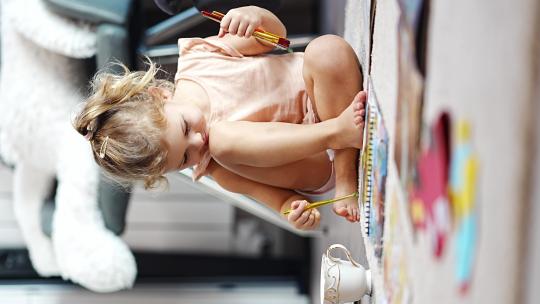 小女孩坐在家里的地板上，用颜料和画笔在一本涂色书上画画