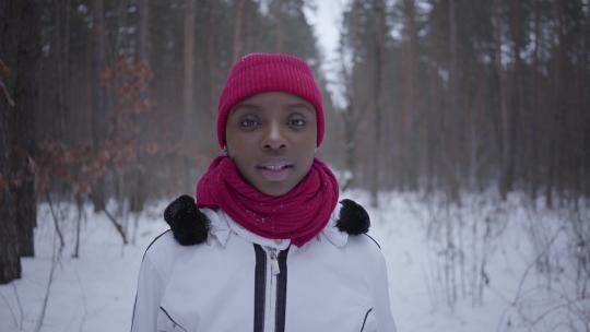 非裔美国女孩走在冬季森林里看着相机，然后起来。穿着保暖夹克的美丽女孩在户外度过时光。慢动作拍摄