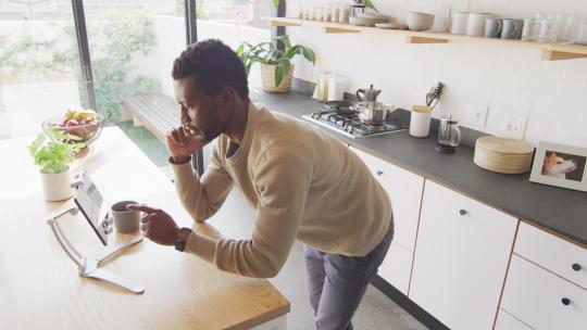 快乐的非裔美国人靠在厨房的台面上，使用平板电脑，用智能手机交谈