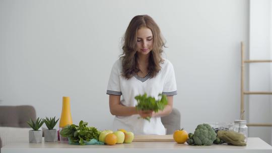 年轻微笑的女人站在现代厨房的餐桌前嗅着新鲜的绿色蔬菜