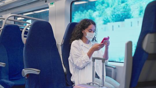年轻的女学生乘坐火车，坐在靠窗的座位上，使用智能手机，戴口罩预防新冠病毒感染，公共铁路交通的乘客，通过手机使用免费wifi