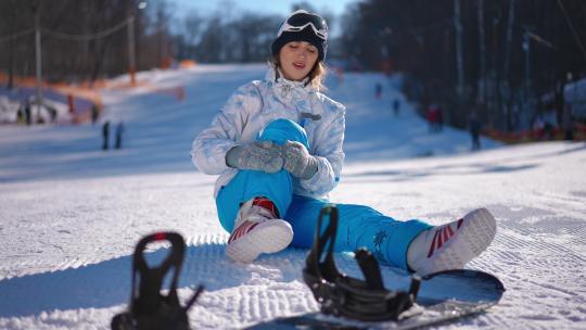 宽镜头年轻女子爱抚受伤的腿坐在白雪上，前面有滑雪板。晴天滑雪胜地膝盖和脚疼痛的美丽高加索女士的正视图。