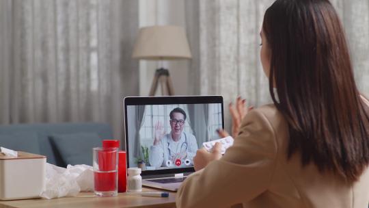 生病的亚洲女商人在工作时拿着药与笔记本电脑上的医生视频通话