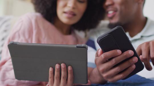 快乐的非裔美国夫妇在客厅使用平板电脑和智能手机