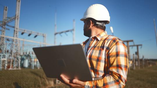 戴头盔的工程师在建筑工地手里拿着笔记本电脑，估算投资。戴安全帽的男工人站在变电站附近