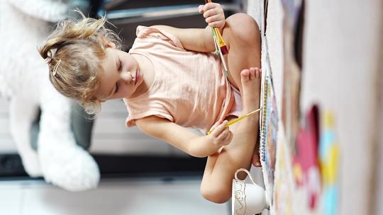 小女孩坐在家里的地板上，用颜料和画笔在一本涂色书上画画