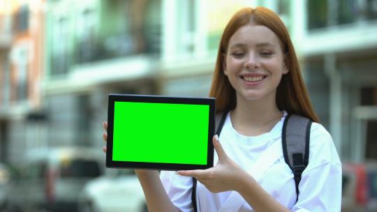 微笑的女孩带着背包显示绿屏平板电脑，廉价旅游和航班视频素材模板下载