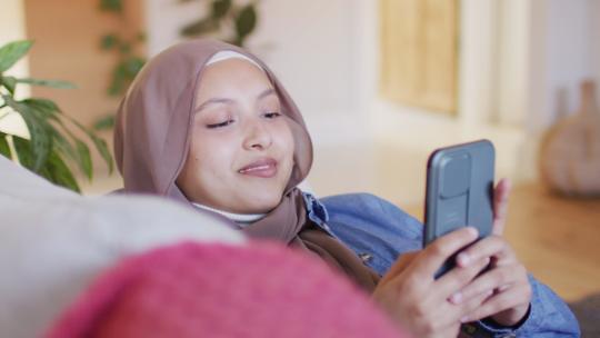 戴头巾微笑的女人在沙发上使用手机