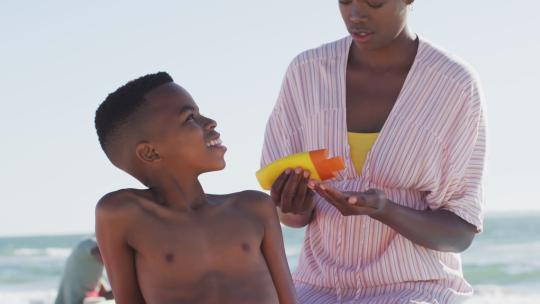 快乐的非裔美国母亲在海滩上给儿子涂防晒霜的视频