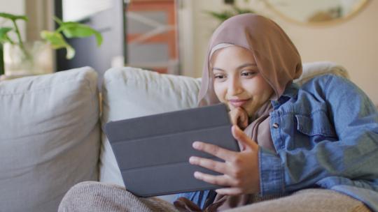 戴头巾的快乐混血儿女人在家里客厅的沙发上使用平板电脑放松的视频