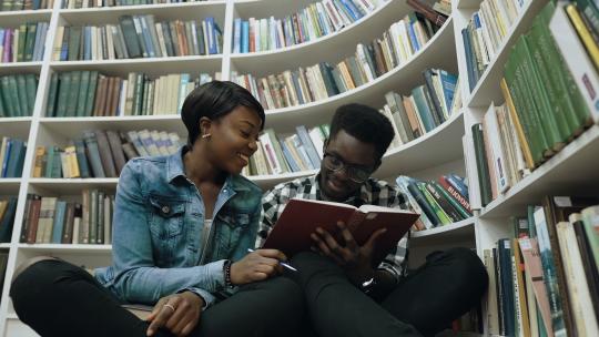 多莉拍摄了年轻的非洲男女学生在现代大学图书馆看书时坐在白色书架附近的地板上。