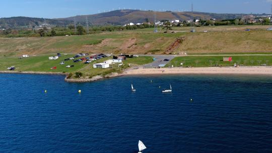 人们在湖边的帆船学校练习，那里有海滩、停车场和风力涡轮机