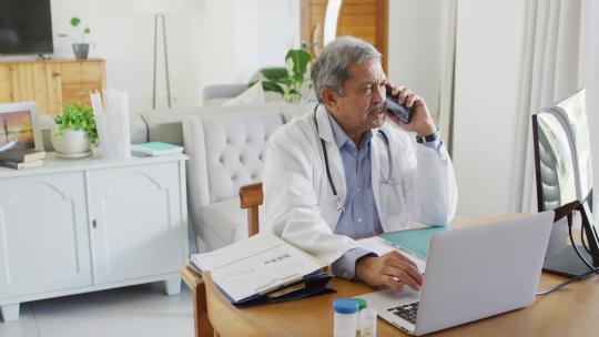 高级混血儿男医生坐在办公桌前用智能手机和笔记本电脑说话