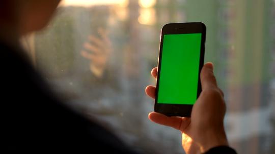 男性手持绿屏智能手机。站在窗户附近使用手机的男子。后视拍摄。色度键