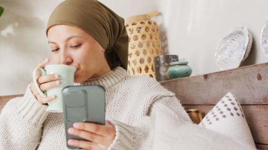 戴头巾的快乐混血女人在家喝咖啡和使用智能手机的视频