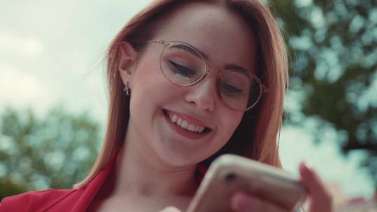 可爱微笑的年轻女子的特写镜头，她像眼镜一样站在户外，积极地发短信，对着屏幕微笑。浏览互联网，社交活跃，博客，instagram。