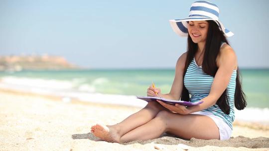 阳光下女人坐在沙滩上拿着笔写作