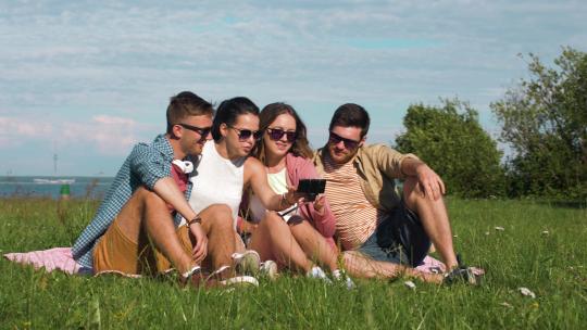 友谊、休闲和科技概念——一群快乐微笑的朋友，带着智能手机和自拍杆在海边或夏季公园户外查看照片