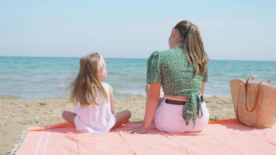 阳光明媚的一天，一对母女背靠背坐在海边沙滩上的橙色毛巾上