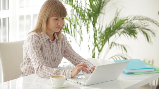 漂亮的年轻女商人坐在桌旁，用笔记本电脑工作，在笔记本上写作