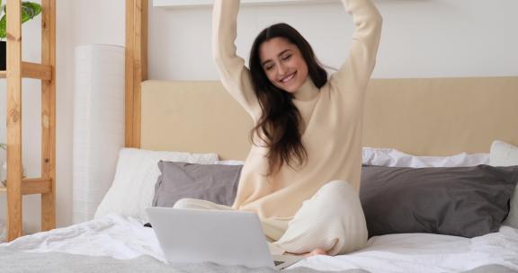 快乐的年轻女子坐在床上，在笔记本电脑前跳舞