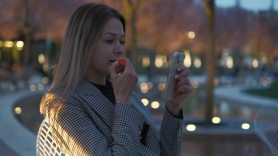 年轻女子约会前在晚上的公园用智能手机作为镜子涂口红