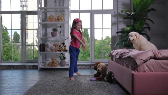 可爱的青春期前女孩穿着睡衣在地毯上为金毛猎犬宠物跳舞，在托儿所的床上聚精会神地观看表演。可爱的小学生表演舞蹈结束屈膝礼