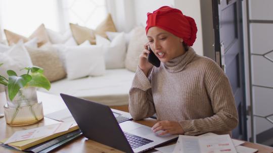 戴着头巾在办公桌上工作的微笑的混血女人在家用智能手机交谈的视频视频素材模板下载