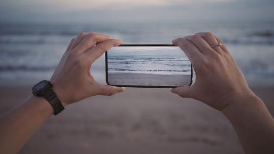 女性手握智能手机新一代oled屏幕的电影屏幕，为社交媒体网络制作照片或视频，海浪不断循环移动。旅行世界的灵感