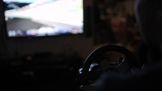 一名男子在黑暗的房间里使用方向盘模拟器玩赛车游戏的特写镜头。在大屏幕上显示速度游戏动作