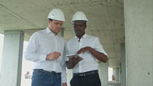 非裔美国工程师和高加索建筑师在建筑工地使用数字平板电脑并佩戴安全帽