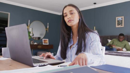 混血女性使用笔记本电脑和阅读笔记的视频，在卧室工作，背景是伴侣