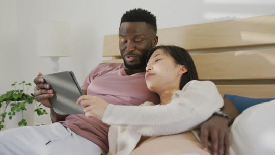 快乐的多样化夫妇使用平板电脑，躺在卧室里