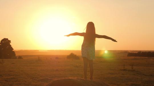 日落时，金色田野里的小女孩站在一卷稻草上，幸福地举起双手。美丽快乐的小女孩剪影在日落的天空中享受户外大自然。