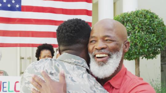 非裔美国男士兵拥抱他微笑的父亲，拥抱家人和美国国旗