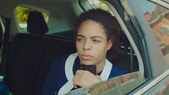 沉思的非裔美国女商人的肖像坐在汽车后座上，用智能手机支撑着下巴，看着车窗外，思考新的商业项目。
