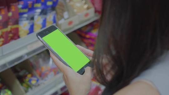女人在超市货架前使用绿屏手机查询商品价格