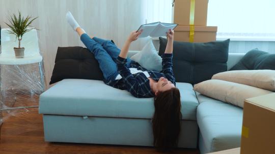 年轻美丽的高加索女人躺在新公寓客厅的沙发上看书