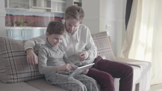爸爸和儿子坐在大客厅的沙发上使用数字平板电脑。父母和孩子之间的关系。快乐的家庭在一起度过时光。爸爸教他的孩子。父子关系。