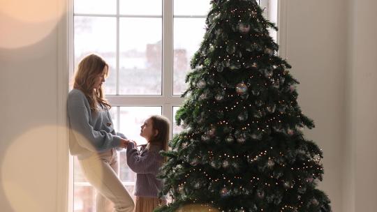 圣诞树前景上妈妈和女儿站在窗前牵手