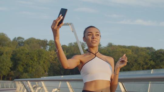 美丽的运动非裔美国妇女在慢跑后为她的vlog在手机上直播视频，在桥上行走时，在清晨旭日的光芒中展示美丽的城市景观。