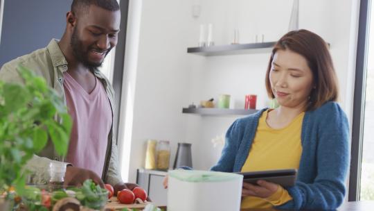 快乐的多样化夫妇在厨房准备食物、使用平板电脑和堆肥蔬菜的视频