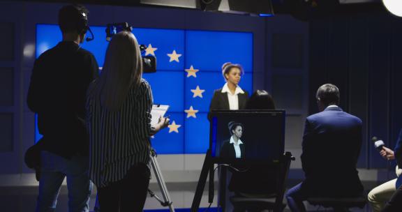 女新闻主播在演播室与制作团队和摄影师一起在欧洲国旗前阅读新闻