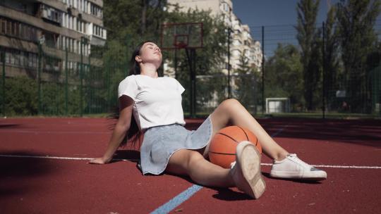 可爱的黑发少女拿着篮球看着坐在篮球场上的相机视频素材模板下载