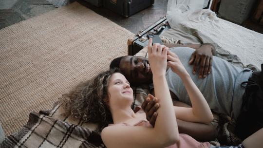 多民族夫妇躺在床上。男人和女人笑着，用智能手机。男性和女性一起度过时光。