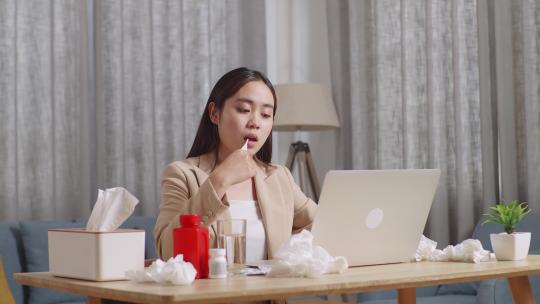 生病的亚洲女商人在使用笔记本电脑工作时使用温度计测试流感