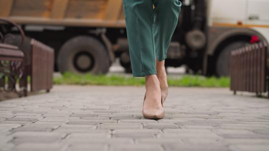 穿着高跟鞋的时尚女人走在铺砌的石板路上，对面是站在路上的自卸卡车。穿着绿色西装的优雅女士去工作特写慢动作