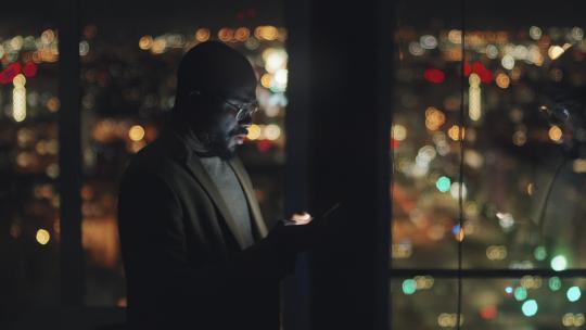 午夜时分，穿着西装、戴着眼镜的非裔美国商人站在黑暗办公室的窗边，揉着额头，使用智能手机，背景是明亮的散景城市景观